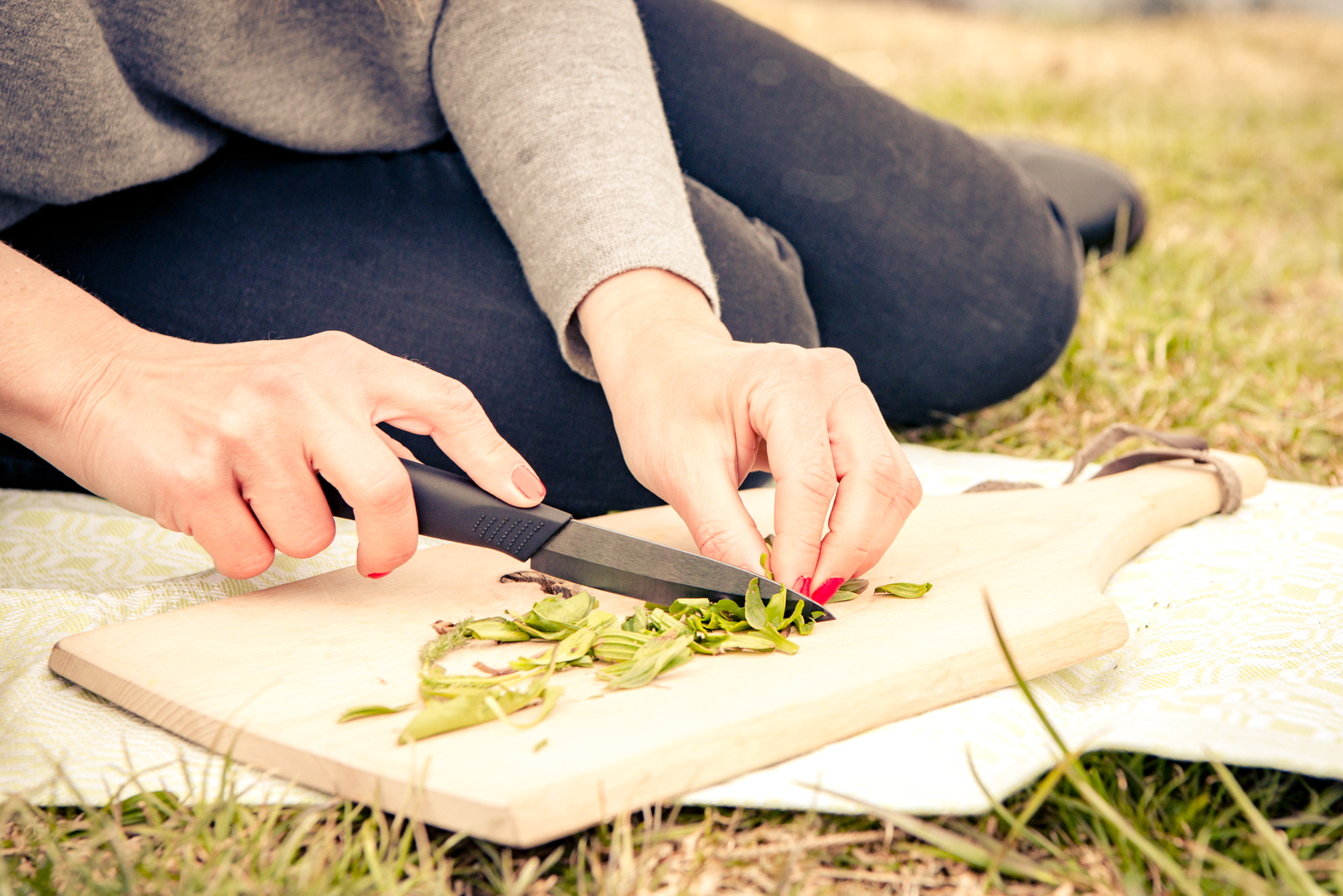 Person, die im Gras sitzt und mit einem schwarzen Messer grüne Kräuter auf einem hölzernen Schneidebrett hackt.