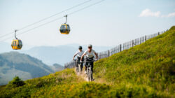 Zwei Personen fahren Mountainbike auf einem grasbewachsenen Hügel mit Seilbahnen und Bergen im Hintergrund.