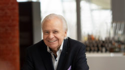 Älterer Mann im dunklen Anzug lächelt - Eckard Witzigmann sitzt in einem Haus, moderner unscharfer Hintergrund.