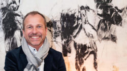 Peter Husty, ein lächelnder Mann mit einem Schal, sitzt vor einem abstrakten schwarz-weißen Pferdebild.