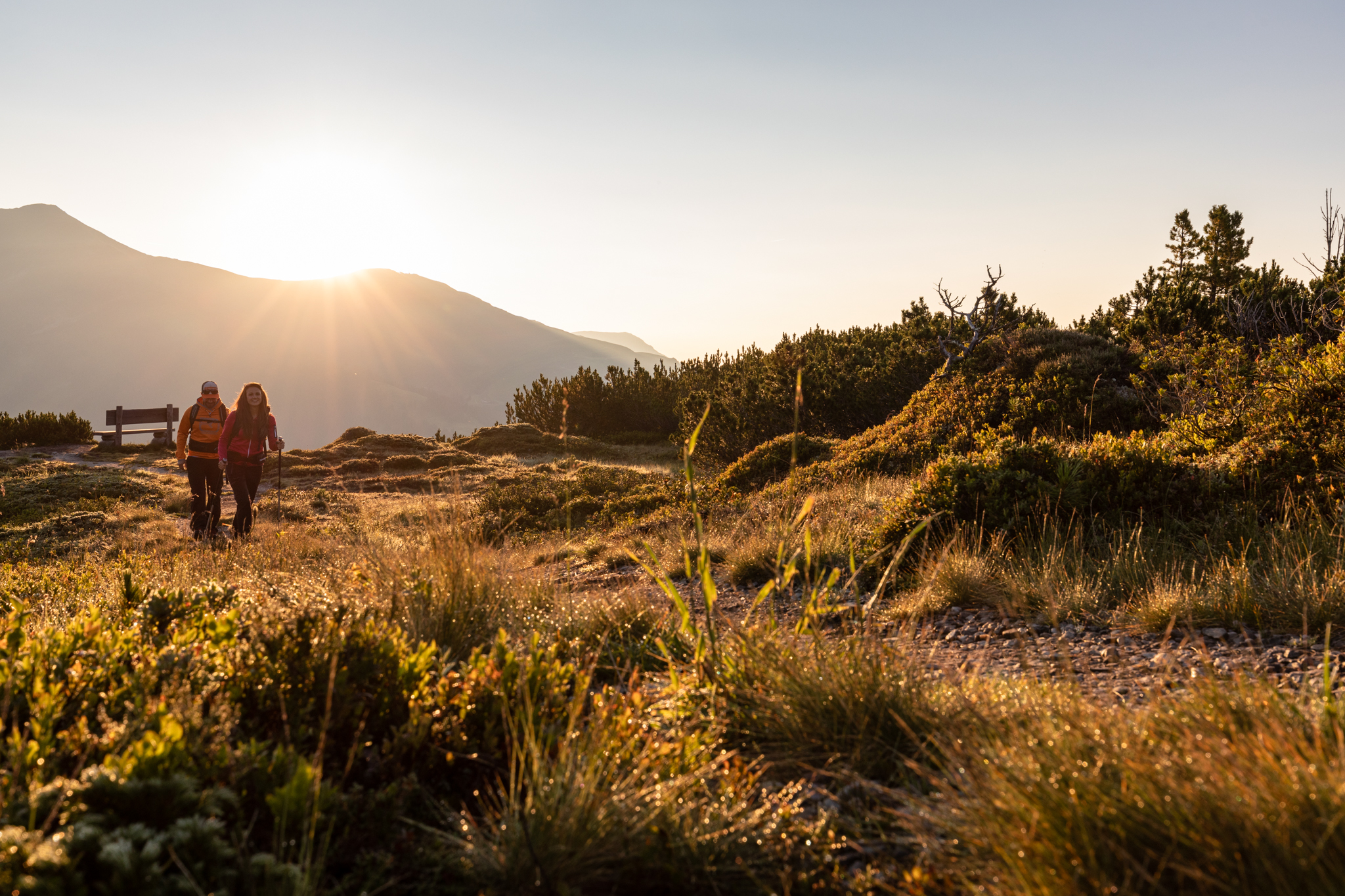 Zwei Personen wandern bei Sonnenuntergang über einen grasbewachsenen Weg mit Bergen im Hintergrund.