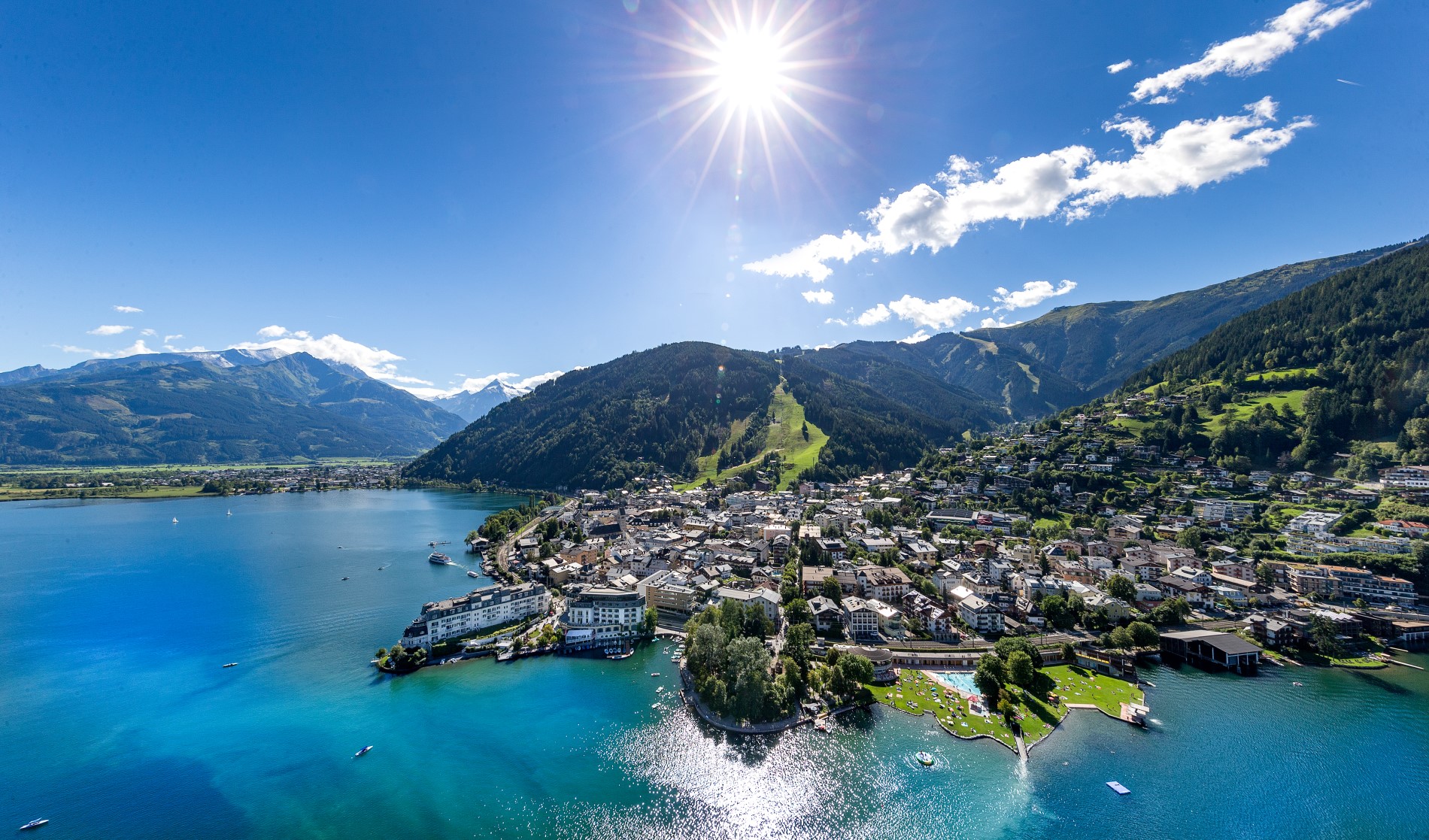 Ein sonniger See, umgeben von Bergen und einer Stadt am Hang, unter strahlend blauem Himmel.