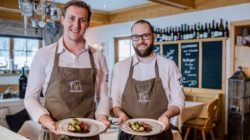 Zwei lächelnde Kellner in Schürzen, wie Florian Zillner, servieren die Speisen in einem gemütlichen Restaurant mit Holzdekor.