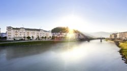 Fluss bei Sonnenaufgang mit Gebäuden, einer Brücke und einem Hügel im Hintergrund unter einem klaren blauen Himmel.