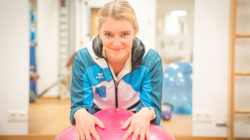 Eine lächelnde Frau in Sportkleidung stützt sich in einem Fitnessstudio auf einen rosa Gymnastikball.