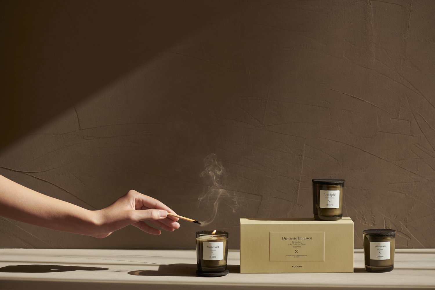 Eine Hand löscht unter anderem eine Kerze und eine beigefarbene Schachtel auf einer hellen Fläche vor einer braunen Wand.