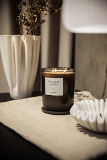 Eine Kerze der Kerzen Manufaktur mit der Aufschrift Nostalgia steht auf einem beigen Tuch neben weißen Vasen und Dekor auf einem Tisch.