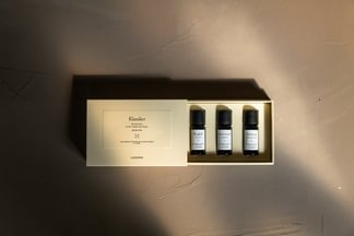 Ein beiger Manufakturkarton mit drei Kerzenflaschen, fein säuberlich angeordnet auf einer braunen Fläche mit sanfter Beleuchtung.