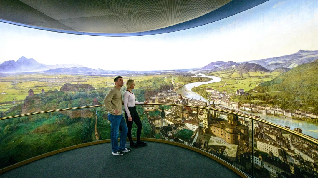 Zwei Personen stehen auf einer Aussichtsplattform und betrachten ein gro&szlig;es Panoramagem&auml;lde mit einer malerischen Landschaft und einer Stadt.