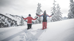 Zwei Personen in Winterkleidung stehen mit ausgestreckten Armen im tiefen Schnee, umgeben von verschneiten Bäumen und Bergen.
