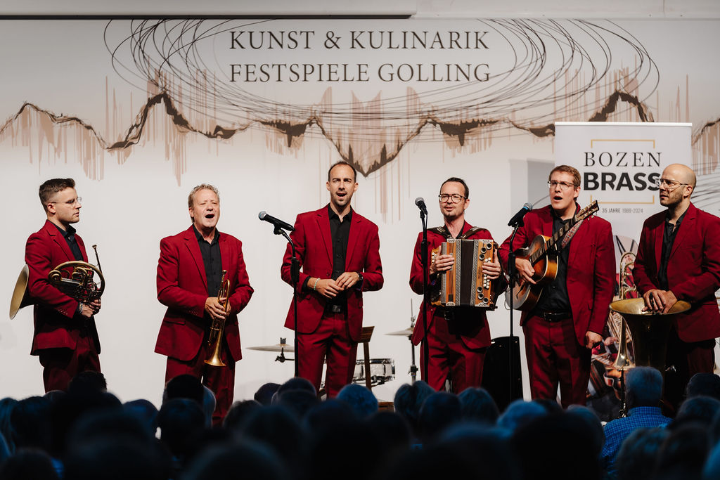 Sechs Musiker in roten Anz&uuml;gen treten mit Blechblas- und Akkordeoninstrumenten bei einem Festival auf.