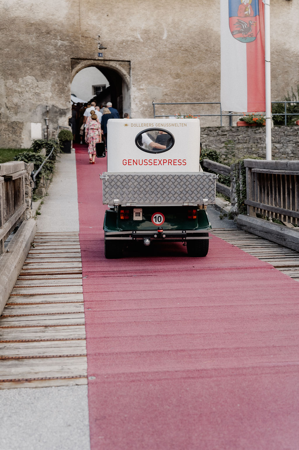 Ein kleines Nutzfahrzeug mit der Aufschrift GENUSS EXPRESS f&auml;hrt auf einem roten Teppich auf einen gew&ouml;lbten Steineingang zu.