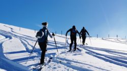Drei Personen genießen einen Skitourenkurs, während sie auf einer verschneiten Piste unter blauem Himmel bergauf fahren.