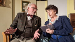 Ein älterer Mann und eine Frau sitzen im Gespräch; die Frau liest aus einem kleinen Notizbuch in der Theatergruppe Koppl.