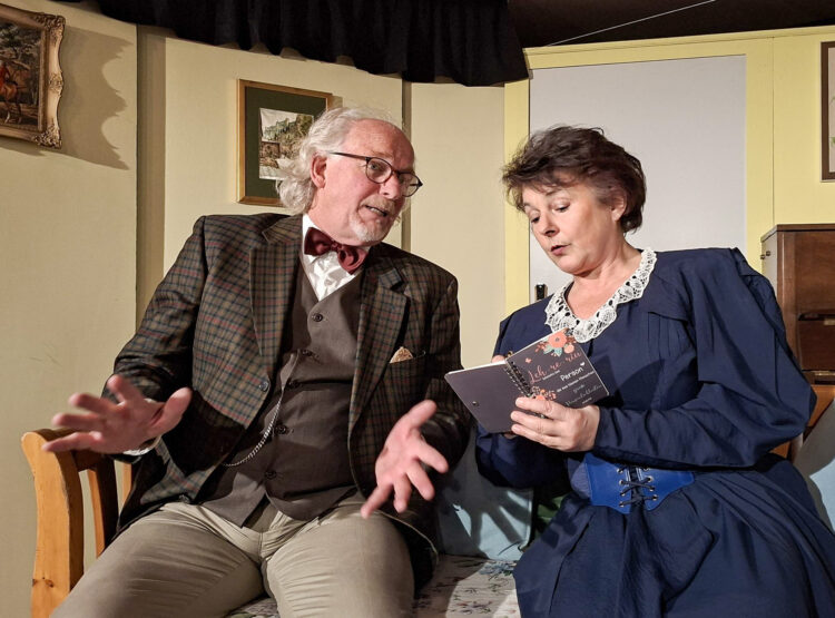 Ein älterer Mann und eine Frau sitzen im Gespräch; die Frau liest aus einem kleinen Notizbuch in der Theatergruppe Koppl.