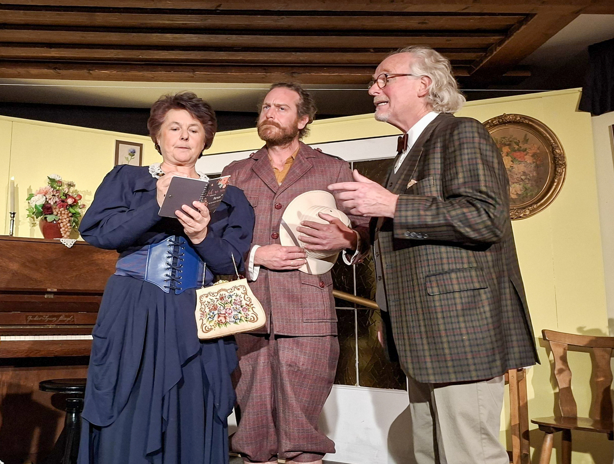 Drei Mitglieder der Theatergruppe Koppl in historischen Kost&uuml;men treten in einem Innenraum auf, w&auml;hrend eine Frau zwei M&auml;nnern aus ihrem Notizbuch vorliest.