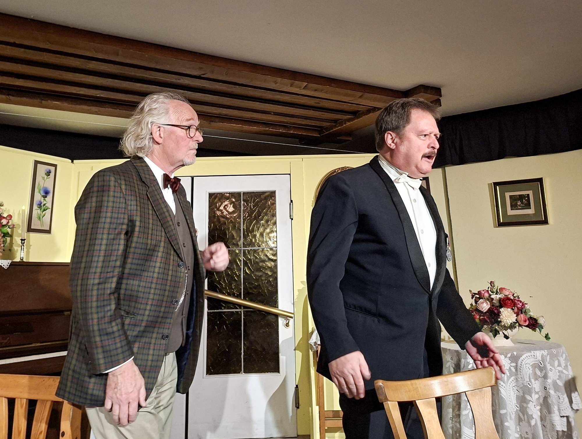 Zwei M&auml;nner in Anz&uuml;gen spielen auf der B&uuml;hne der Theatergruppe Koppl, im Hintergrund sind Blumen und ein Klavier zu sehen.