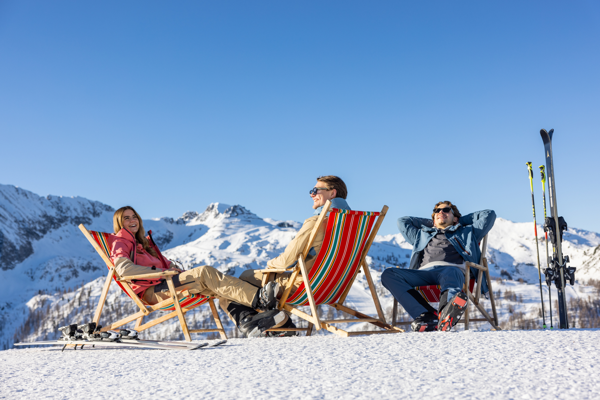 Drei Personen entspannen sich auf Liegestühlen im Schnee mit Skiern in der Nähe, Berge und blauer Himmel im Hintergrund.