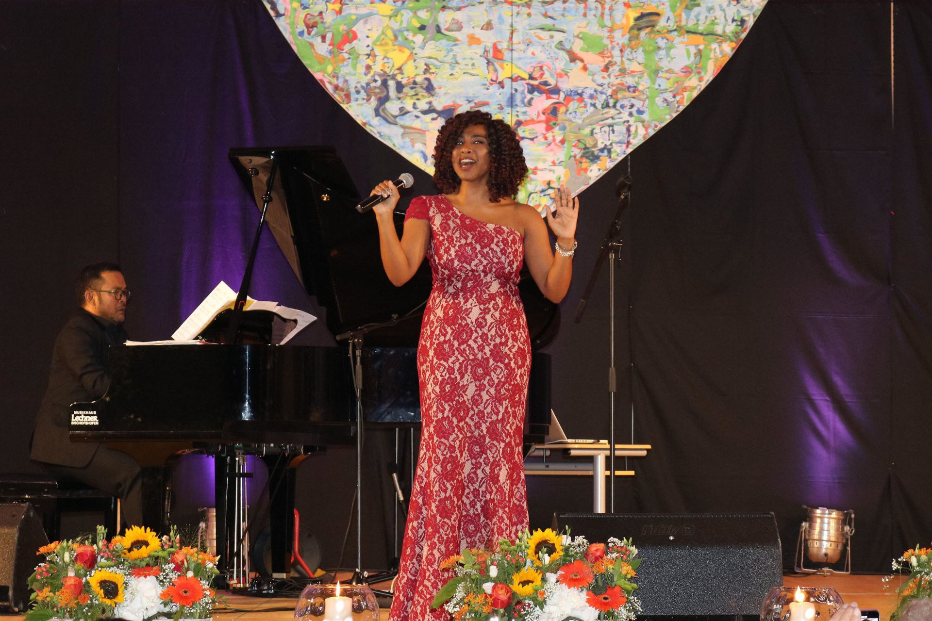 Eine Frau in einem roten Abendkleid singt auf der Bühne, im Vordergrund sind ein Pianist, bunte Blumen und ein UNICEF-Banner zu sehen.