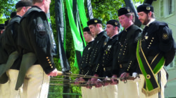 Männer in schwarzen Uniformen mit grünen Schärpen führen einen Schwerttanz auf, wobei sie in Reihen stehen und ihre Schwerter aufeinander richten.