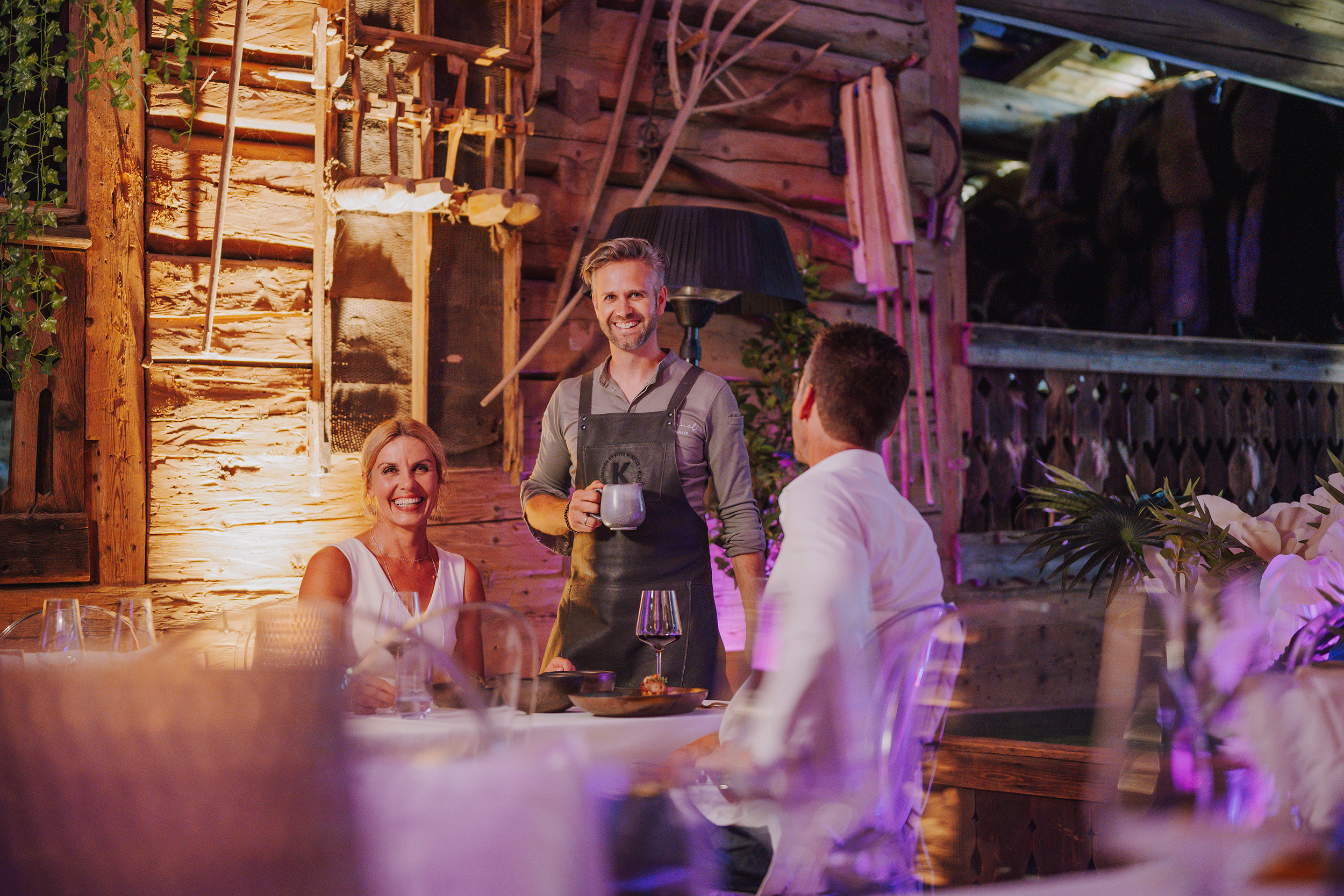 Ein lächelnder Kellner bedient ein glückliches Paar in einem rustikalen, warm beleuchteten Restaurant.