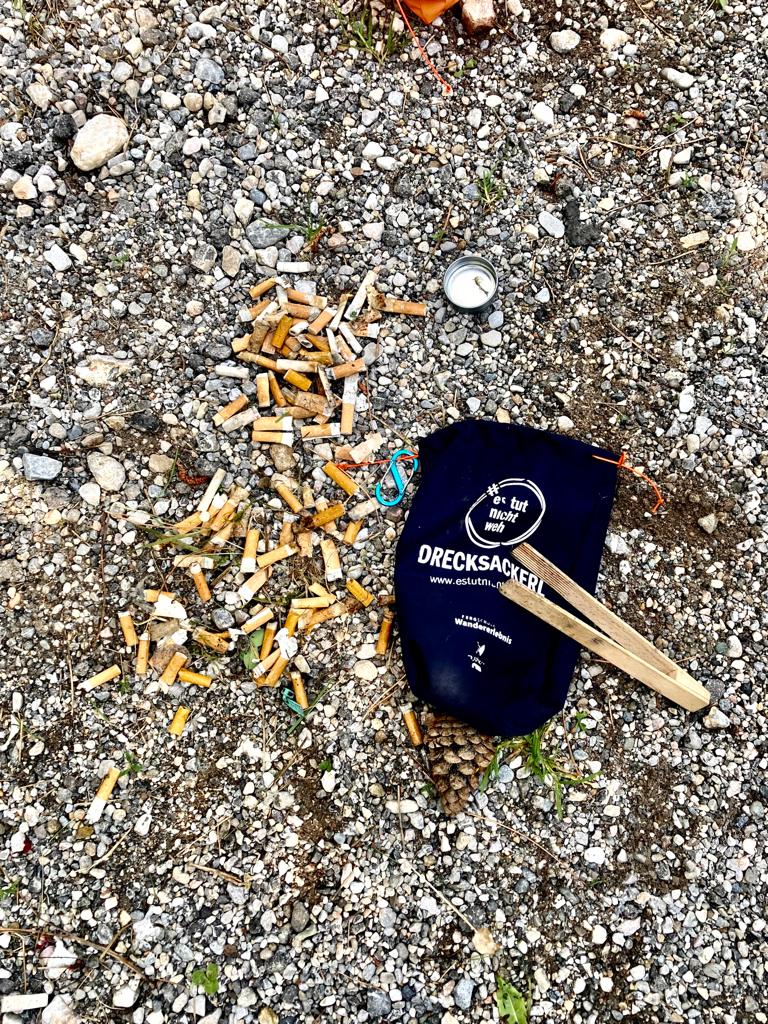 Ein Haufen Zigarettenstummel, ein Beutel, ein Flaschendeckel und eine Zange auf felsigem Boden.