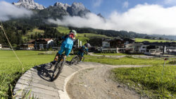 Zwei Mountainbiker fahren auf einem gewundenen Feldweg mit grünen Hügeln und Bergen im Hintergrund.