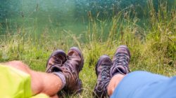Zwei Personen in Wanderschuhen entspannen sich im Gras an einem Gewässer, die Beine zum See hin ausgestreckt.