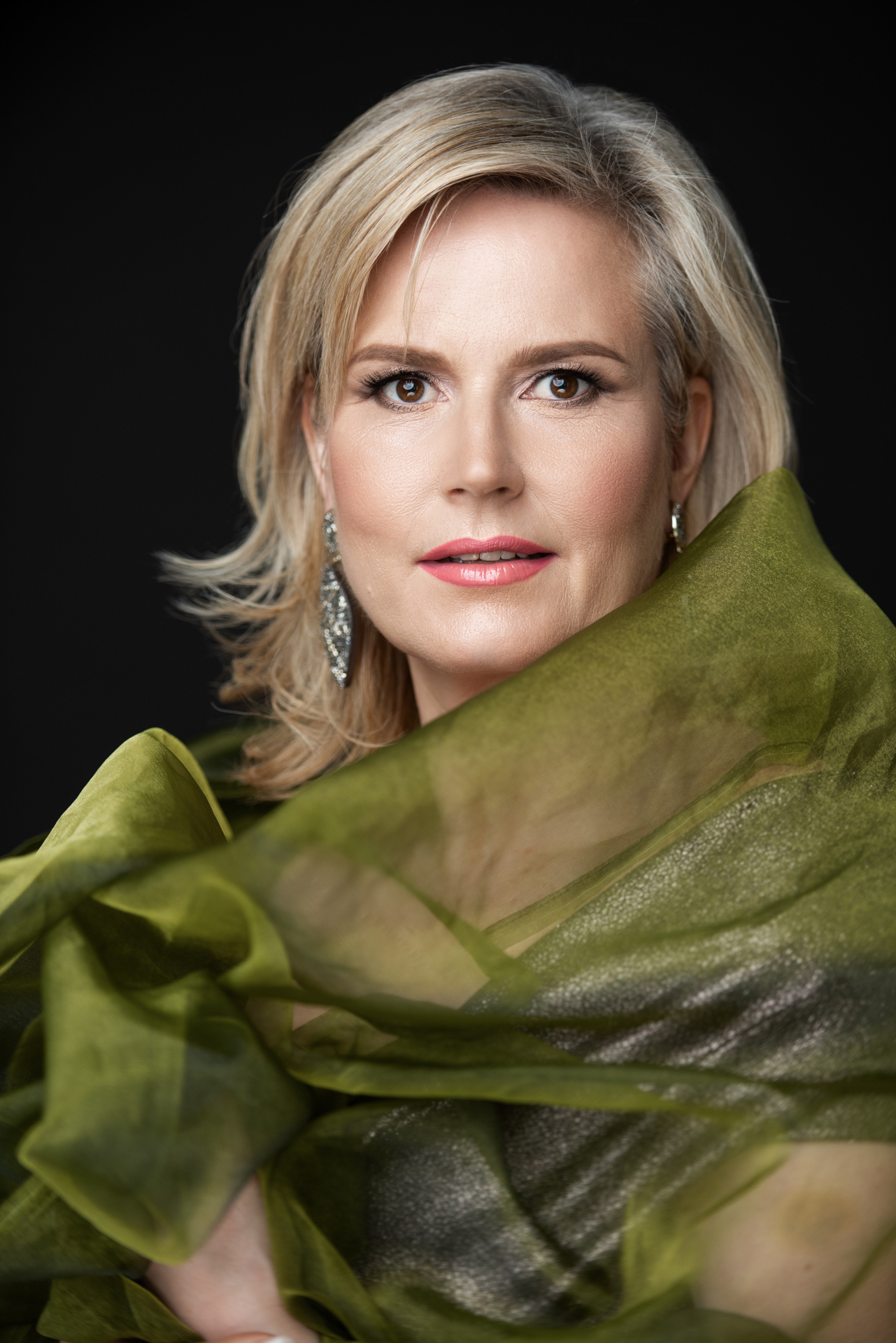 Eine Frau mit blondem Haar und Ohrringen posiert in einem durchsichtigen gr&uuml;nen Stoff vor einem schwarzen Hintergrund, der an Mozarts Eleganz erinnert.