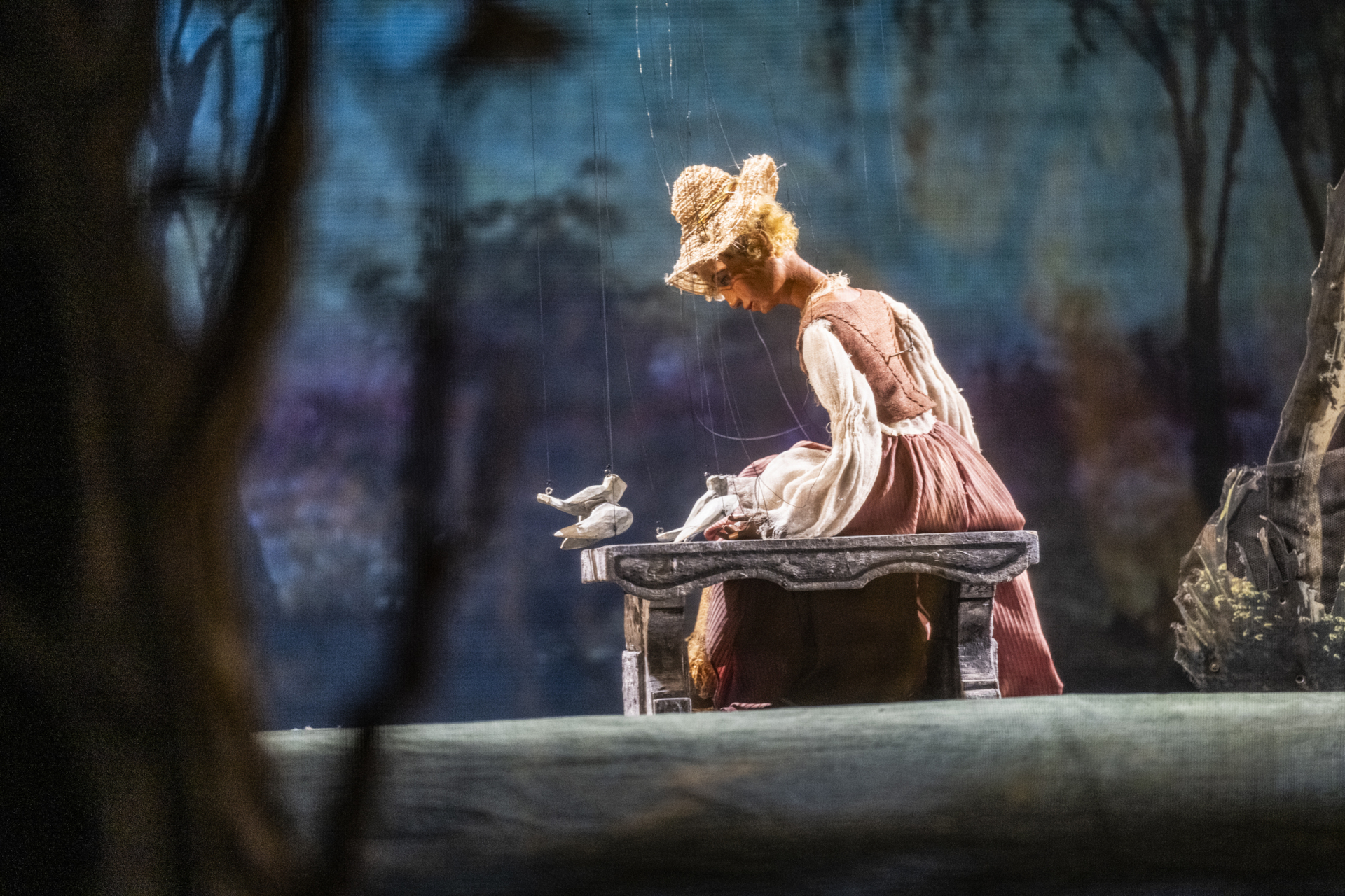 Eine Marionette in Kleid und Haube sitzt auf einer Bank, während Vögel in einer Waldbühne flattern, die den Charme Mozarts versprüht.