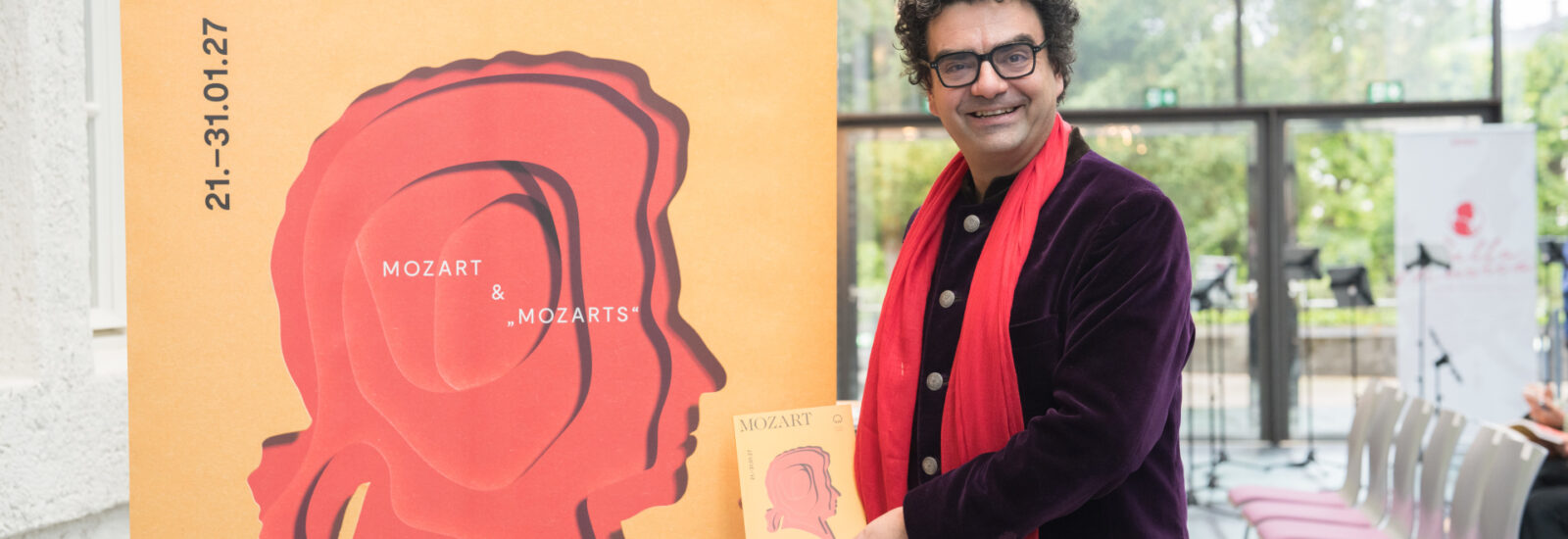 Rolando Villazon mit rotem Schal hält bei einer Indoor-Veranstaltung ein Mozart-Poster neben einem großen Mozart-Display.
