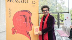 Rolando Villazon mit rotem Schal hält bei einer Indoor-Veranstaltung ein Mozart-Poster neben einem großen Mozart-Display.