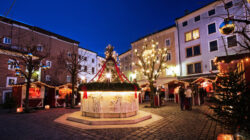 Ein festlicher Weihnachtsmarkt auf einem Stadtplatz mit Lichtern, geschmückten Bäumen und weihnachtlichen Ständen in der Abenddämmerung.