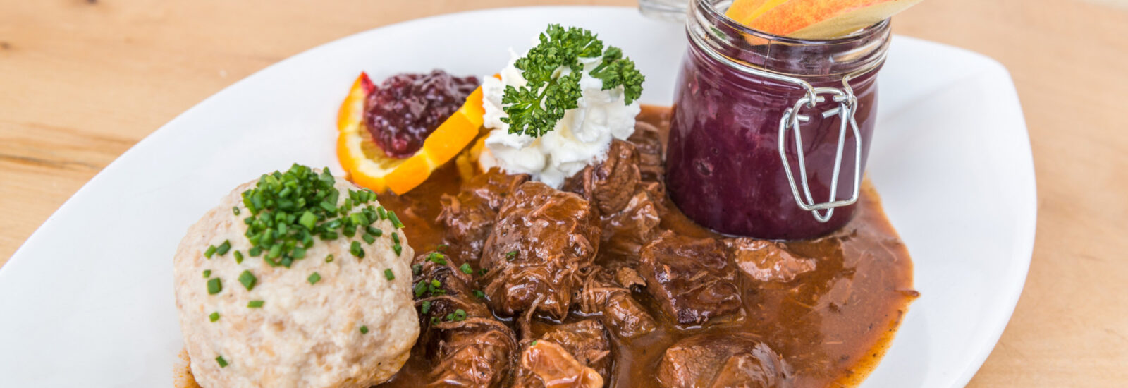Ein Teller mit Rindergulasch, Brotknödel, Rotkohl aus dem Glas sowie Orangen- und Apfelscheiben als Beilage.