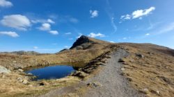 Ein Schotterweg führt auf einen grasbewachsenen Berg mit einem kleinen Teich unter einem blauen Himmel mit vereinzelten Wolken.
