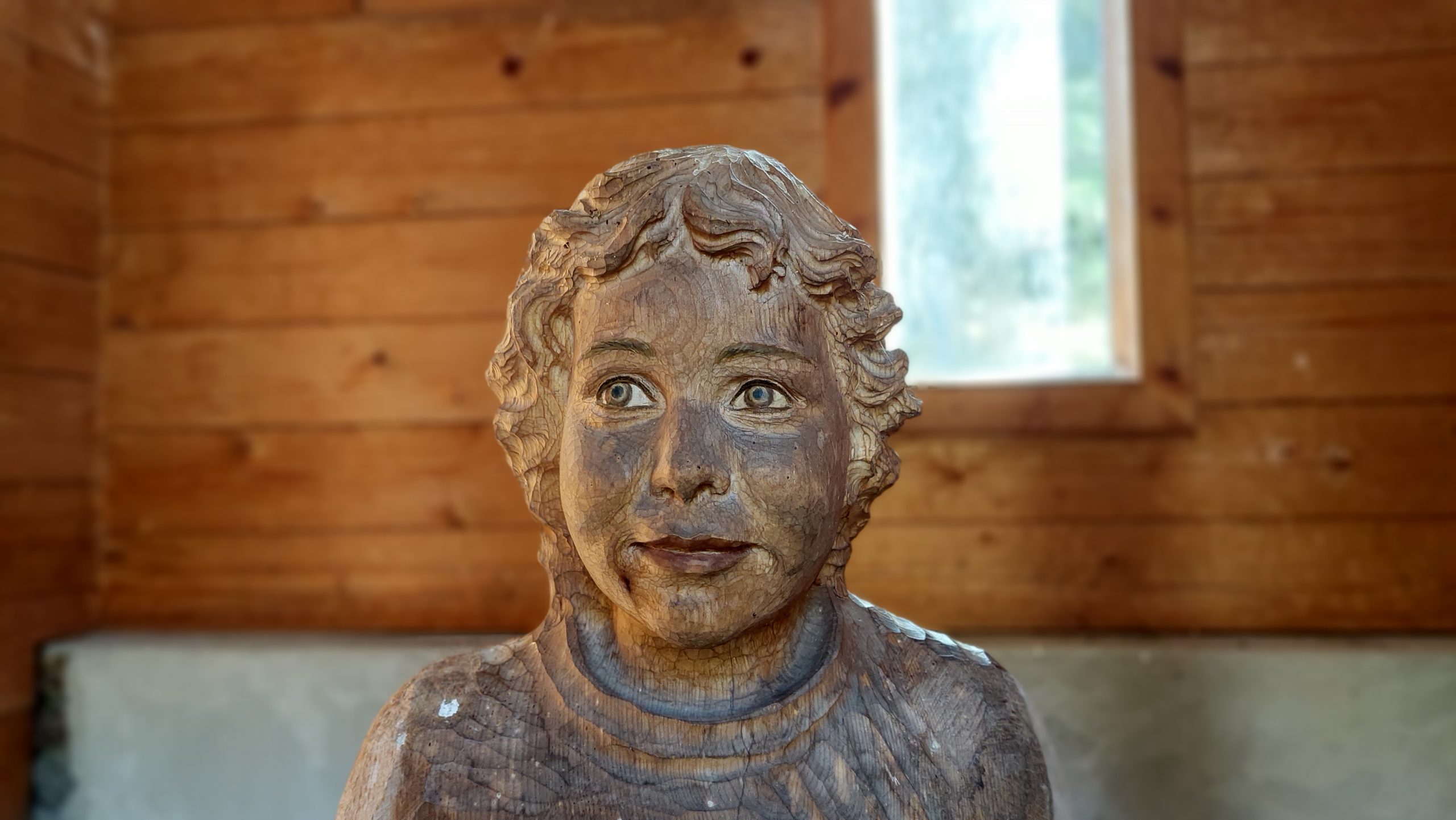 Holzskulptur eines Kindes mit lockigem Haar in einer Holzhütte, mit einem Fenster im Hintergrund.
