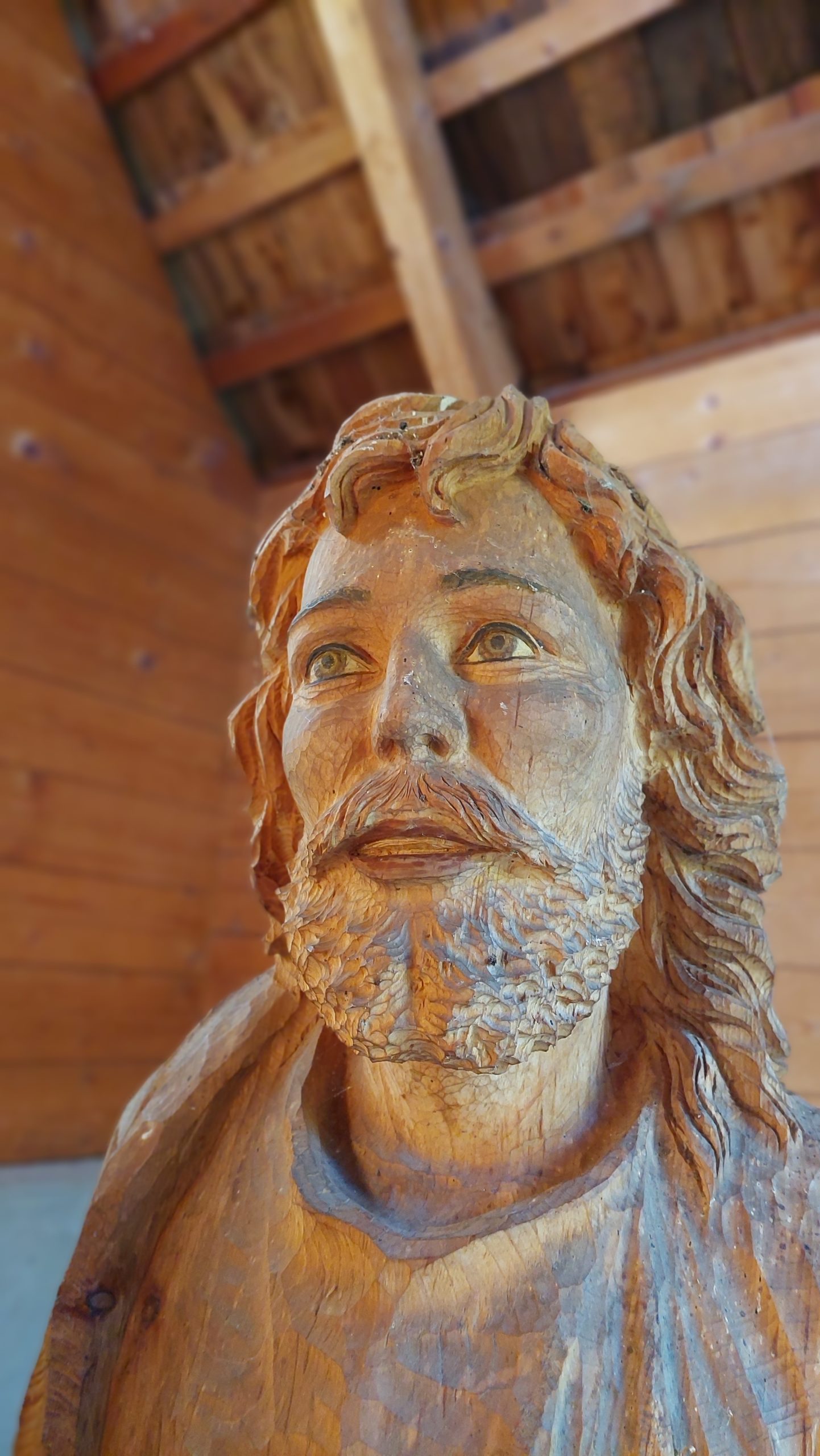 Nahaufnahme einer Holzskulptur, die einen bärtigen Mann mit langen Haaren vor einem hölzernen Hintergrund darstellt.