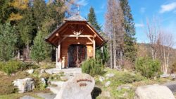 Eine kleine Holzkapelle mit einem dekorativen Tor, umgeben von Bäumen und Felsen unter einem blauen Himmel.
