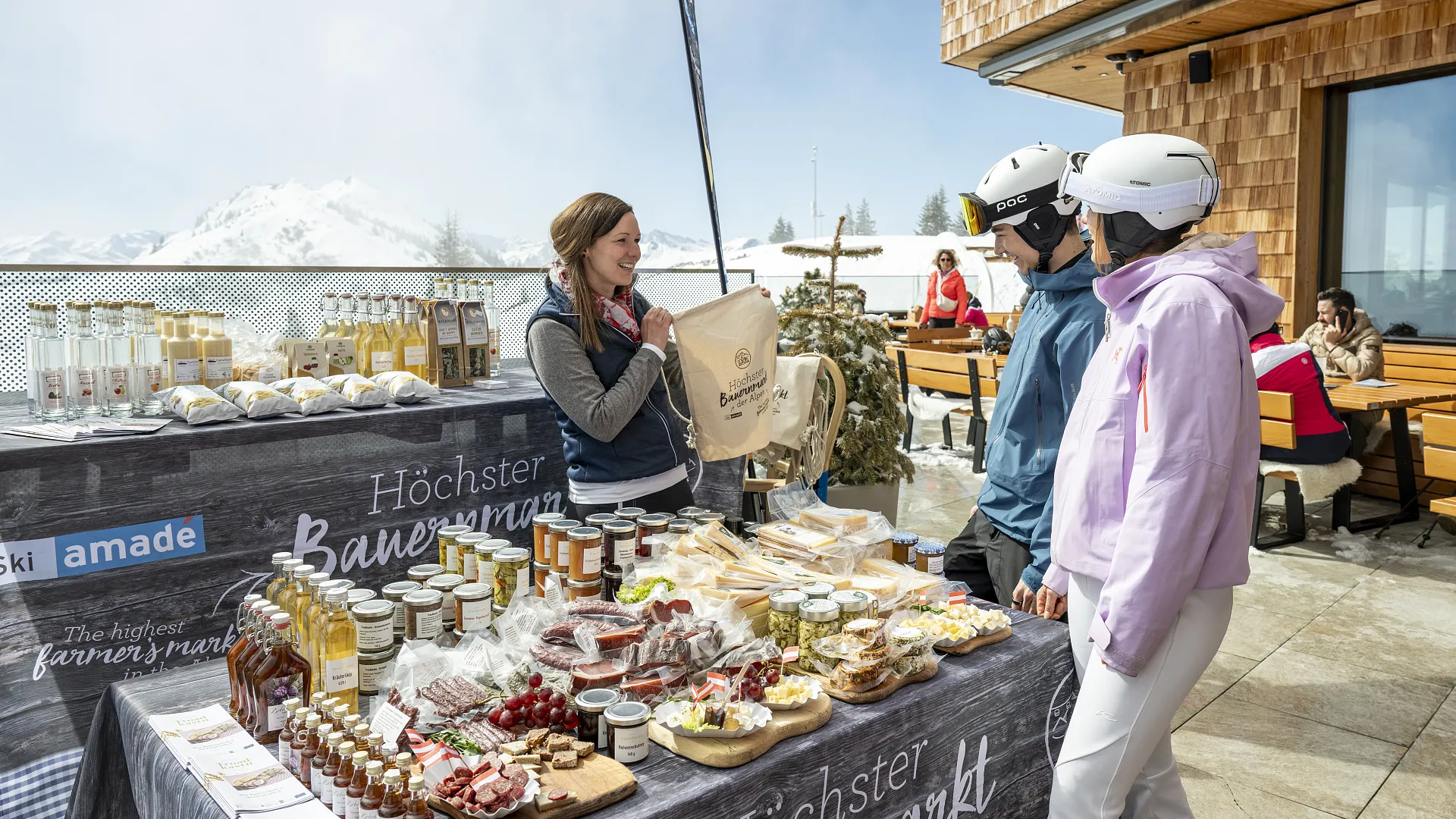 Zwei Skifahrer stöbern an einem Marktstand im Freien in lokalen Speisen und Getränken, während im Hintergrund die verschneiten Berge zu sehen sind.