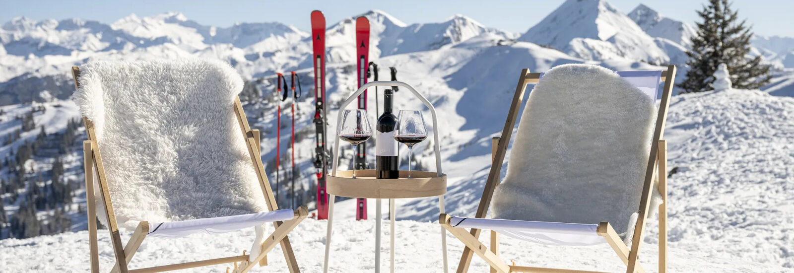 Zwei Liegestühle und ein kleiner Tisch mit Wein auf verschneiten Bergen, mit Skiern und Gipfeln im Hintergrund.