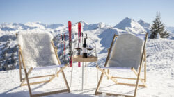 Zwei Liegestühle und ein kleiner Tisch mit Wein auf verschneiten Bergen, mit Skiern und Gipfeln im Hintergrund.