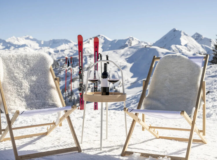 Zwei Liegestühle und ein kleiner Tisch mit Wein auf verschneiten Bergen, mit Skiern und Gipfeln im Hintergrund.