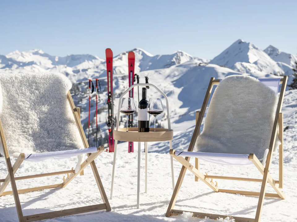 Zwei Liegestühle und ein kleiner Tisch mit Wein auf verschneiten Bergen, mit Skiern und Gipfeln im Hintergrund.