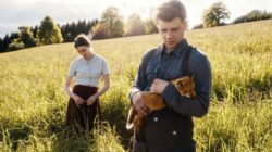 Ein Mann, der ein Fuchsjunges hält, steht in einem sonnigen Feld, hinter ihm eine Frau in Bluse und Rock.