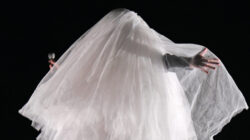 Eine Person, die als Geist in ein weißes Laken gehüllt ist und ein Mikrofon in der Hand hält, steht mit ausgestreckten Armen in der Dunkelheit.