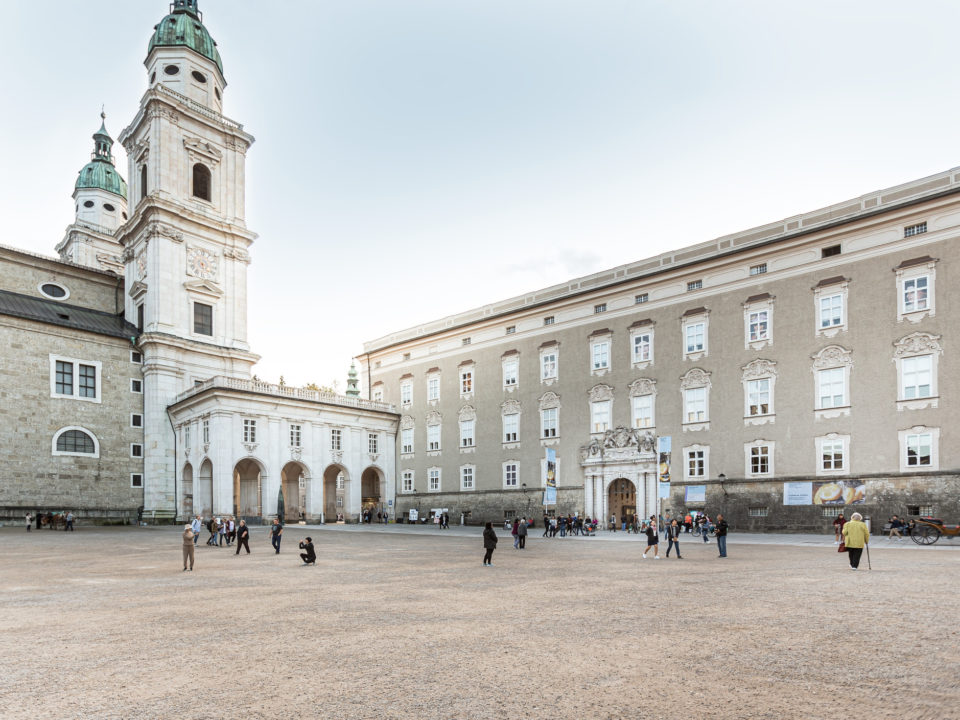 Menschen, die über einen weitläufigen Platz spazieren, der von historischen Gebäuden umgeben ist, darunter eine Kirche mit hohen Türmen.