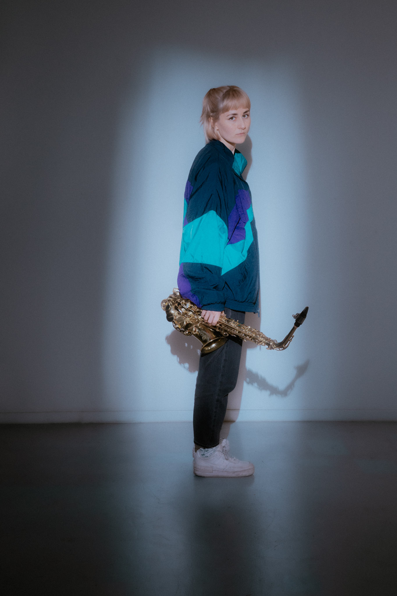 Eine Person in einer bunten Jacke h&auml;lt ein Saxophon in der Hand, das von einem Scheinwerfer in einem schummrigen Raum angestrahlt wird.