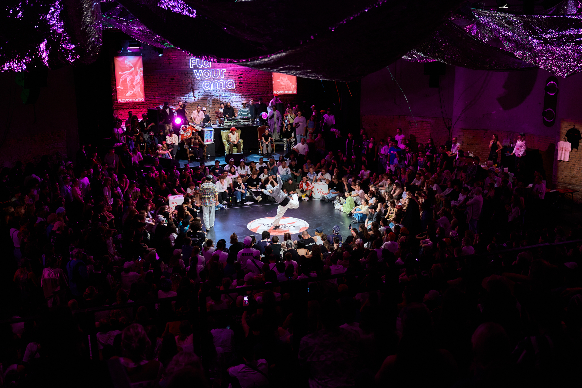 Eine große Menschenmenge verfolgt einen Breakdance-Wettbewerb in einem schwach beleuchteten Saal mit violetter Beleuchtung.