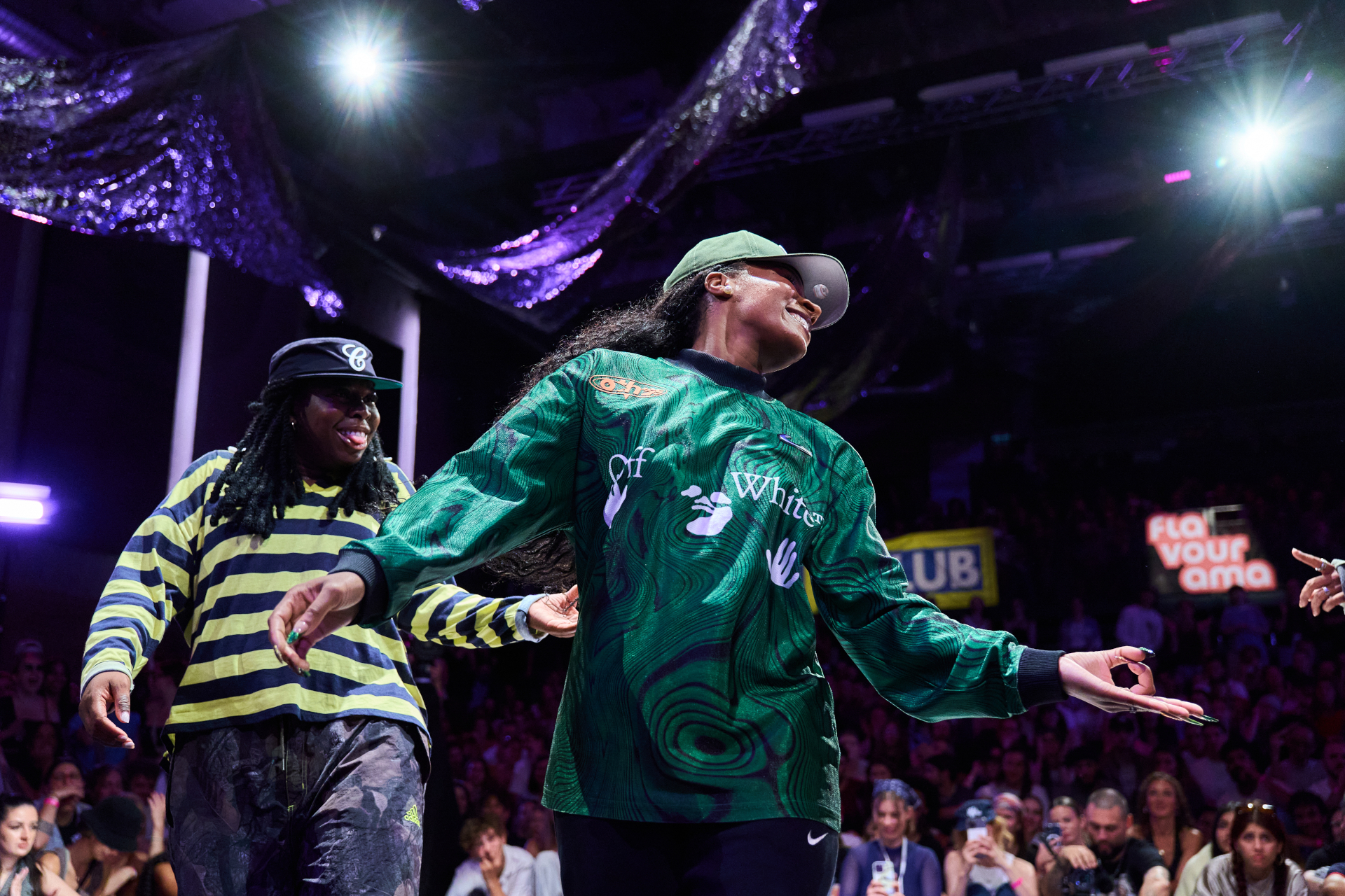 Zwei Tänzerinnen in farbenfrohen Kostümen treten auf der Bühne vor einem jubelnden Publikum energisch auf.