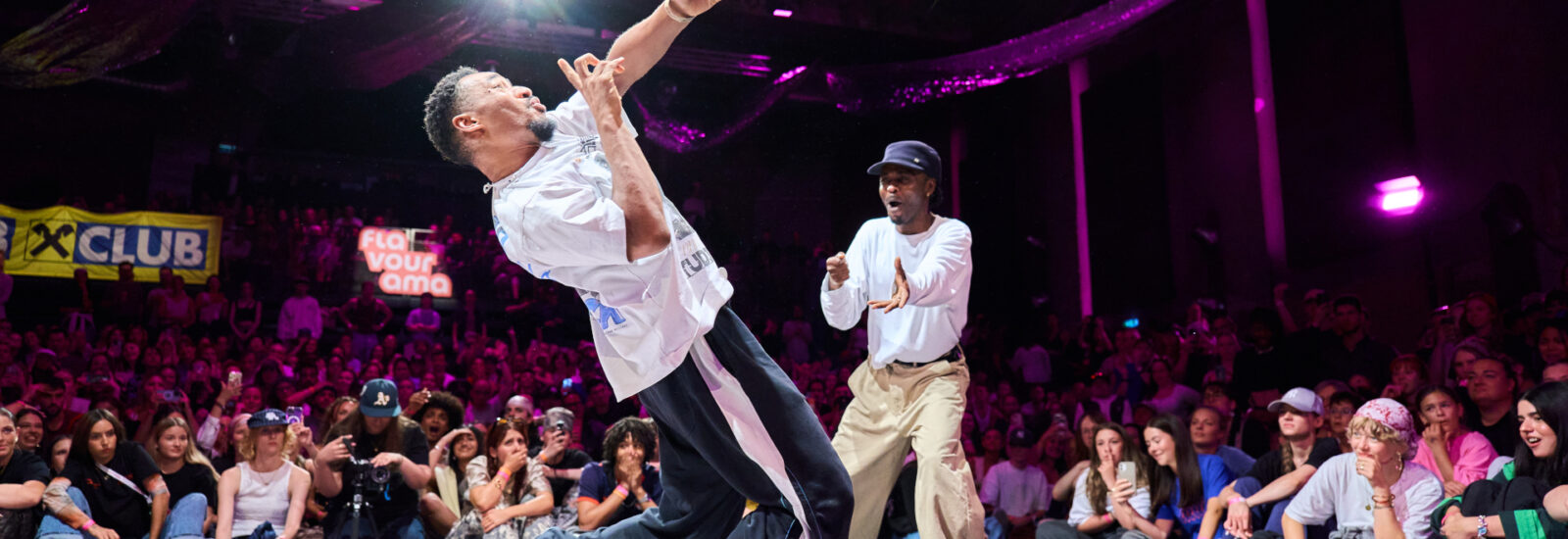 Zwei Männer breakdancen auf der Bühne vor einer jubelnden Menge und bunten Lichtern im Hintergrund.